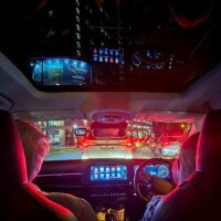 Car interior at night with sun shade open. View of dahsboard lit in red and blue vibrant colors. A tall building appears in the sun roof. Picture taken from back seat. Showing driver and front passenger half image.