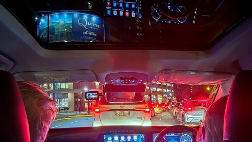 Car interior at night with sun shade open. View of dahsboard lit in red and blue vibrant colors. A tall building appears in the sun roof. Picture taken from back seat. Showing driver and front passenger half image.